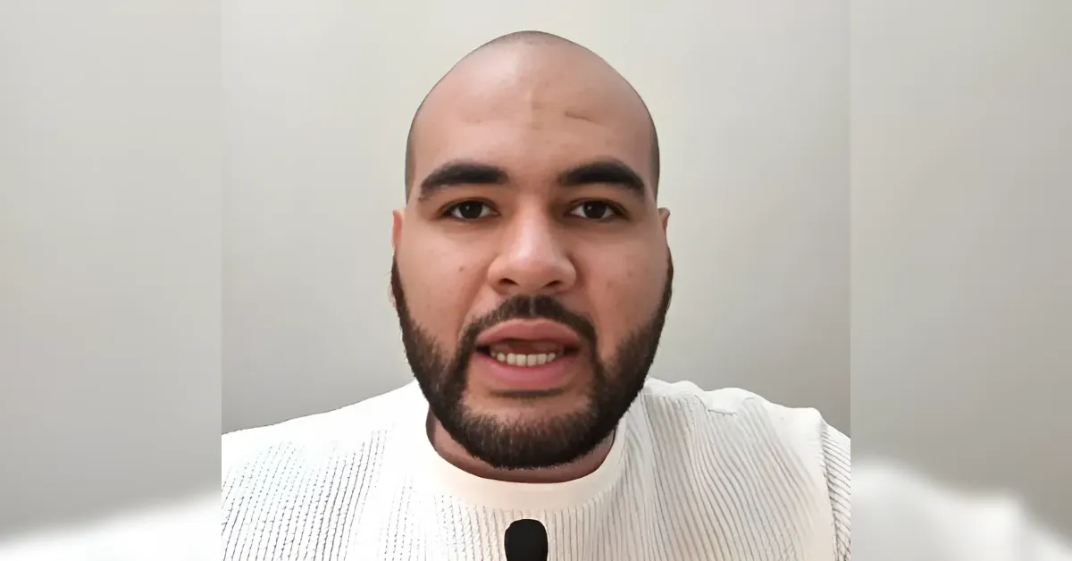 Man with a beard speaking directly to the camera, wearing a white knit sweater against a neutral gray background.