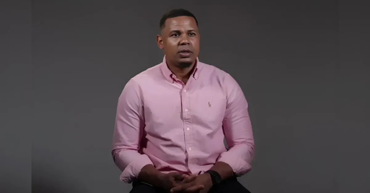 Man in a pink button-down shirt sits against a gray studio backdrop, speaking to the camera.