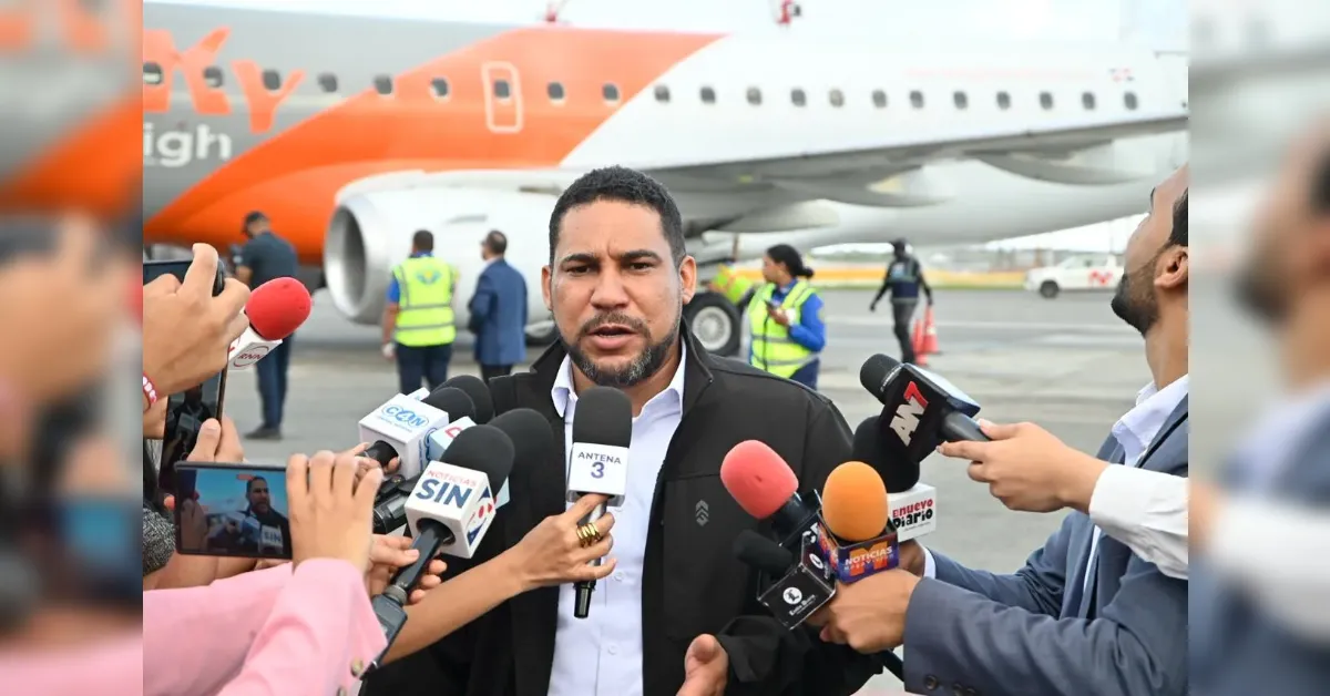 Man in a dark suit speaks to a cluster of reporters holding microphones at an airport tarmac, with an orange airplane behind him.