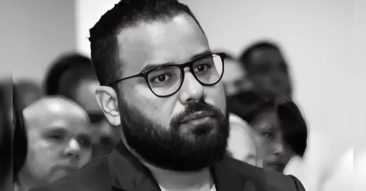 Man with black-rimmed glasses and beard among a blurred crowd at an event (black and white).