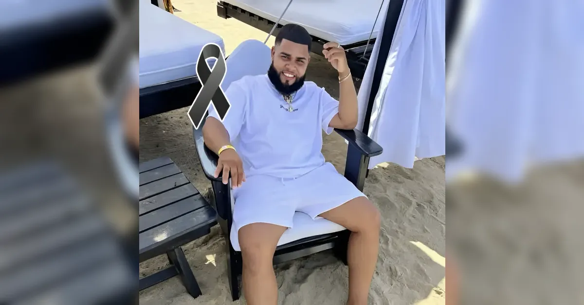 Man in a white outfit sits in a beach chair on sandy ground, smiling under a shade tent with a black mourning ribbon overlay.