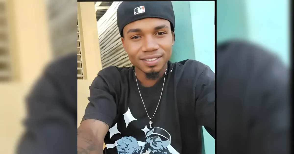 Young man in a black graphic t‑shirt and backward cap smiles at the camera outdoors, wearing a silver chain with a cross.