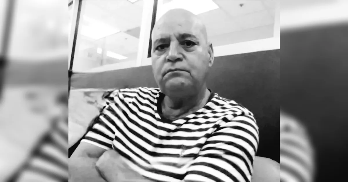Older man with a striped shirt sits with arms crossed, looking at the camera in a grayscale photo.