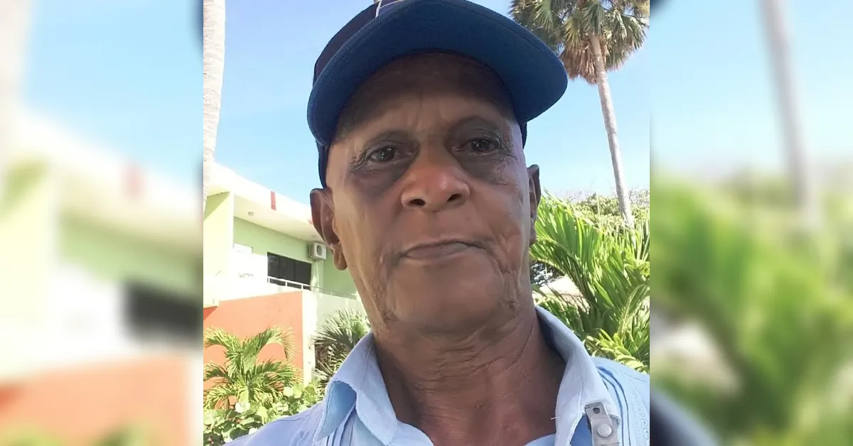 Portrait of an older man in a blue cap and light blue shirt, outdoors with palm trees and a building in the background, sunny day.