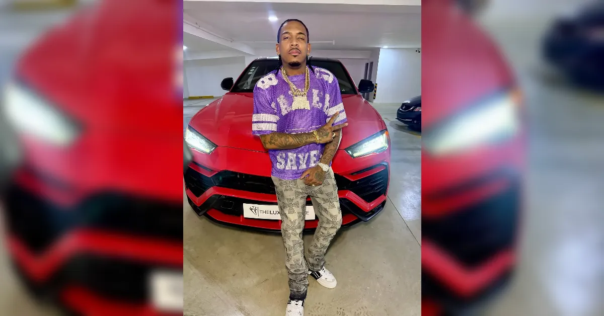 Man wearing a purple graphic t-shirt and patterned gray jeans stands with arms crossed in front of a red car in a parking garage.