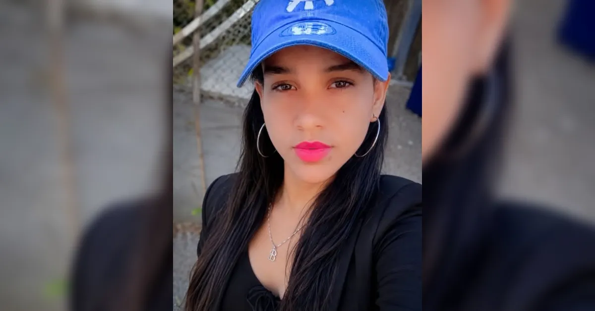 Portrait of a woman outdoors wearing a blue cap, black outfit, large hoop earrings, pink lipstick, and a necklace with a pendant