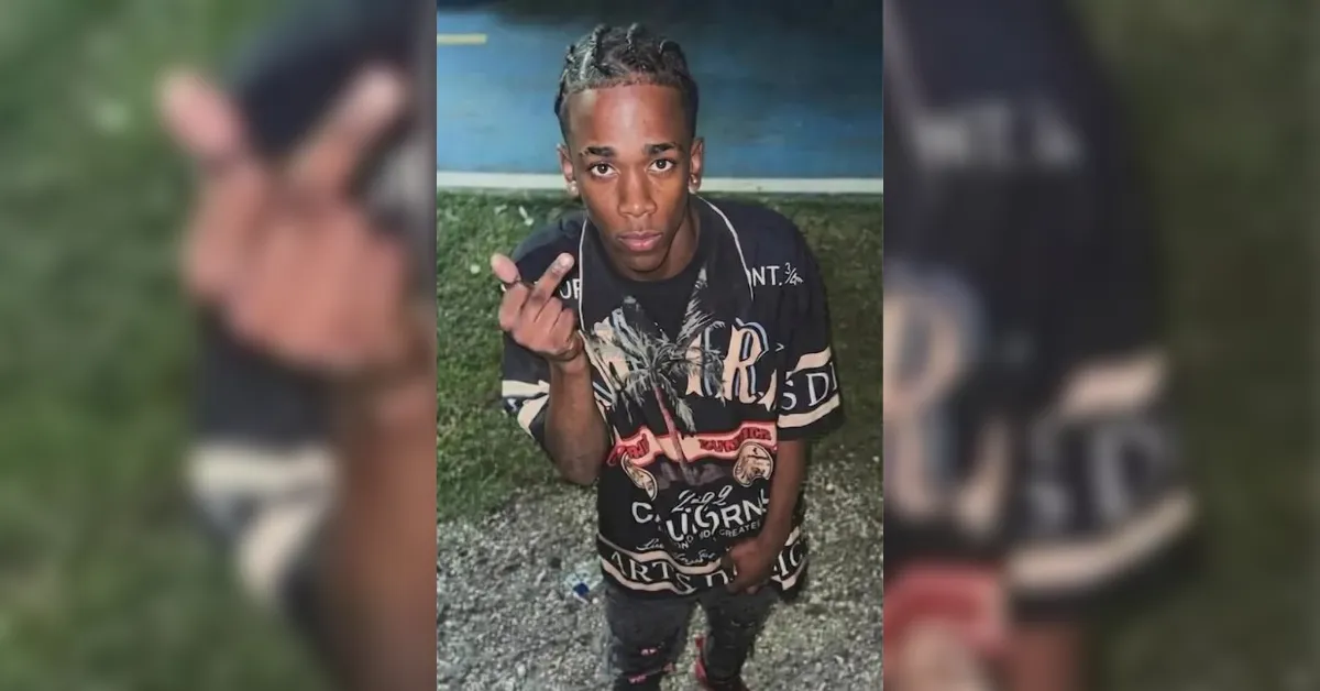 Young man with braided hair stands outdoors on gravel, raises his middle finger at the camera and wears a black graphic T-shirt with designs in white and red.