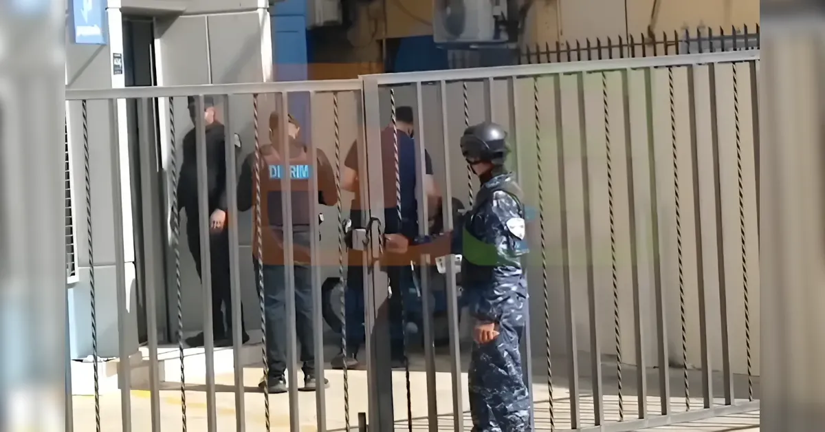 Security officer in camouflage uniform and helmet stands by a tall metal fence as civilians talk near a gated entrance.