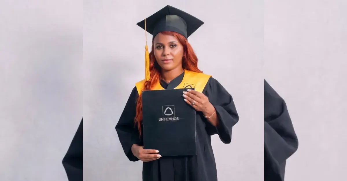 Graduate in a black cap and gown with a yellow stole, holding a diploma cover with a university emblem.