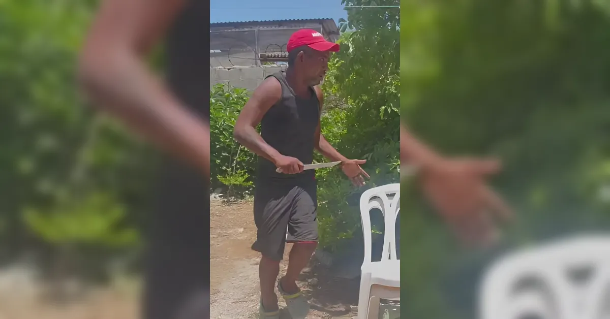 Man in a red cap and black sleeveless shirt holds a large knife while standing outdoors near a white plastic chair.