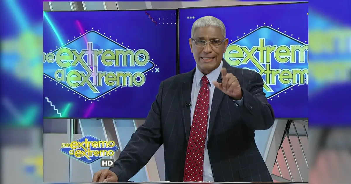 Male TV host in a dark suit and red tie points toward the camera on a colorful studio set with a logo reading 'Extremo' in the background.