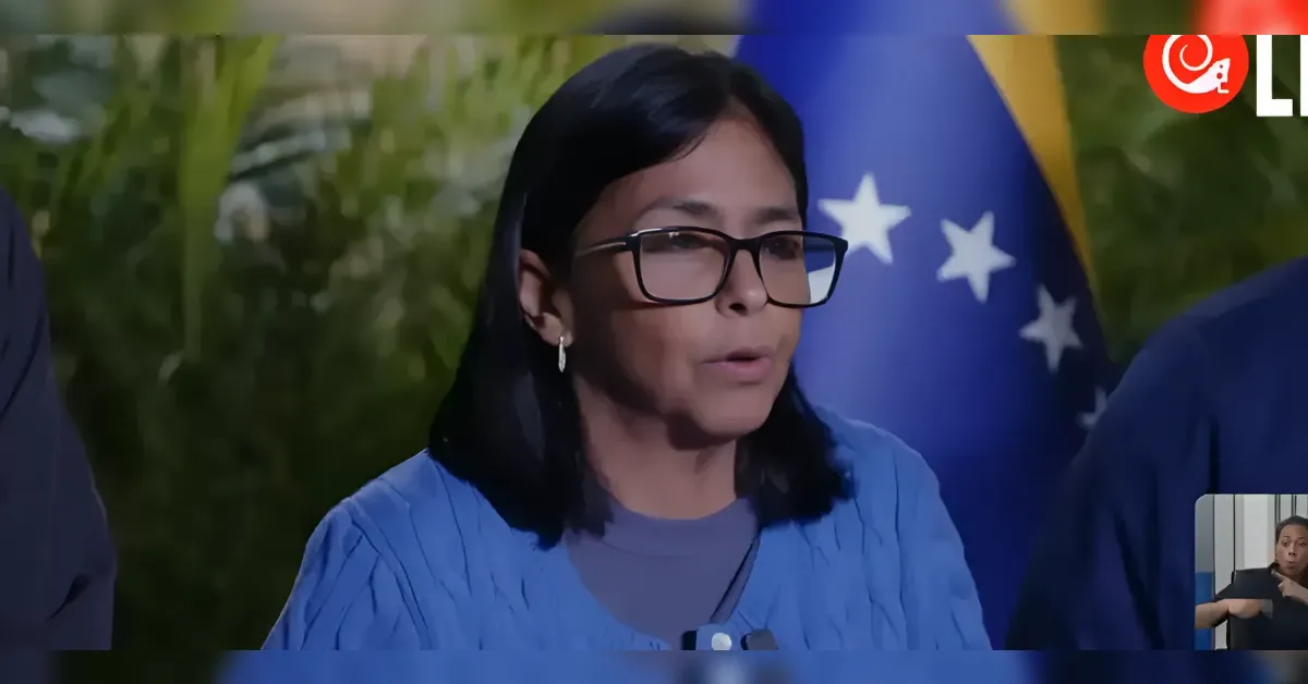 Woman with dark hair and glasses speaks during an interview, blue top, with flags in the background.