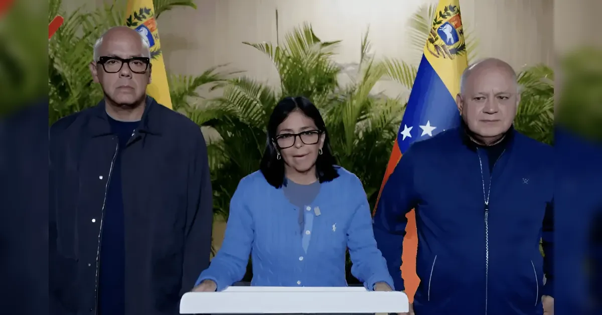 Three people stand behind a podium at a press conference: a woman in a blue cardigan at center, flanked by two men, with a Venezuelan flag in the background.