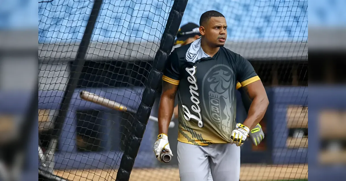 Male baseball player in a black and gold jersey, holding a bat, standing beside a batting cage net.