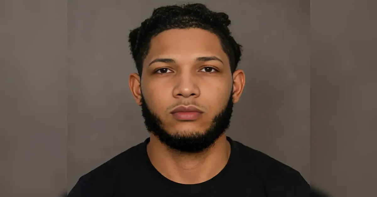 Portrait of a young man with short curly hair and a full beard, wearing a black shirt, looking straight at the camera against a gray background.