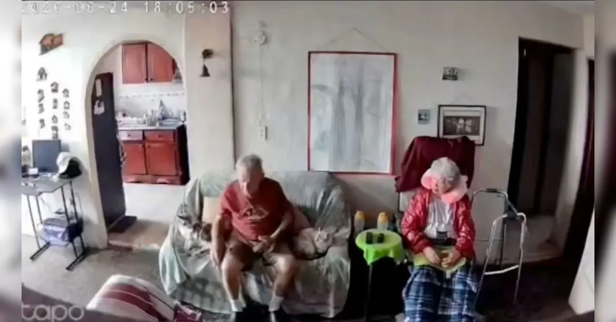 Two elderly people sit on a sofa in a living room, with a walker nearby and a small green table between them; kitchen visible through an arched doorway behind them.