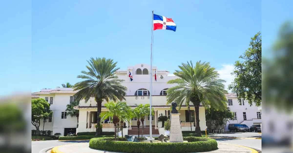 White two-story building with arched windows, palm trees, and a flagpole flying the Dominican Republic flag in a circular driveway area with a bust statue nearby.