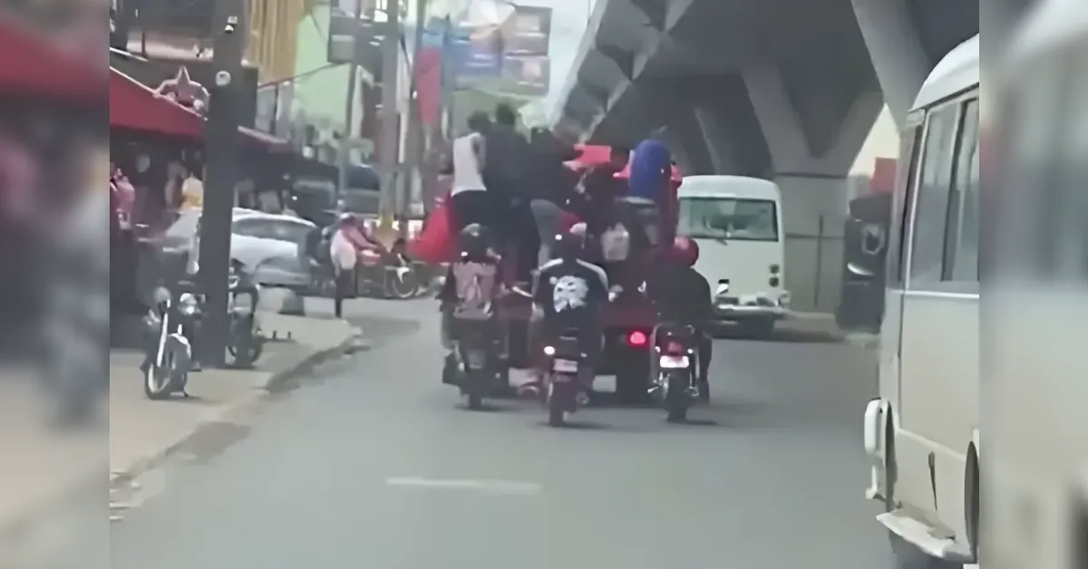 Motorcycle riders and passengers crowded on a city street under an overpass, with a bus ahead and pedestrians nearby.