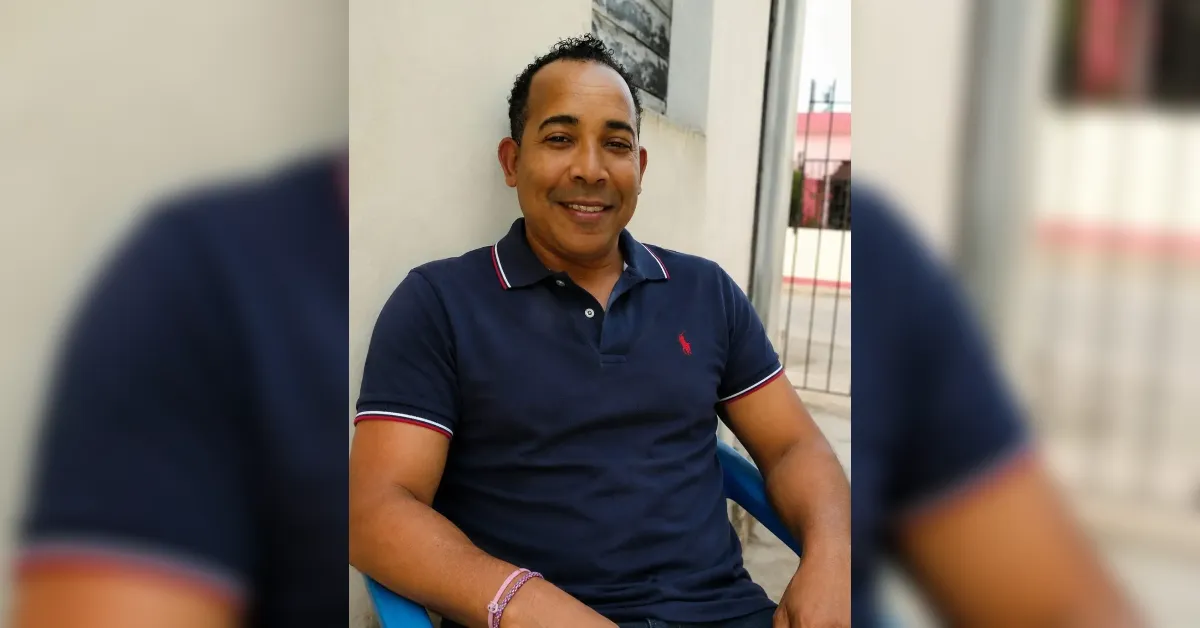 Smiling man in a navy polo shirt sits outdoors against a light wall in a blue chair.