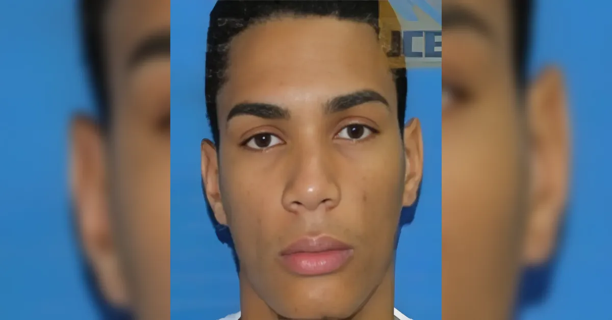 Close-up portrait of a young man with short black hair and a neutral expression against a blue background.