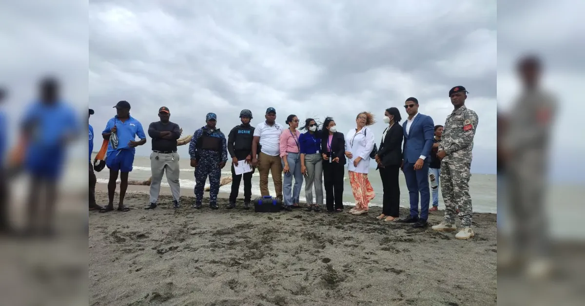 A diverse group of people, in uniforms and professional attire, lined up on a sandy beach under a cloudy sky for a group photo.