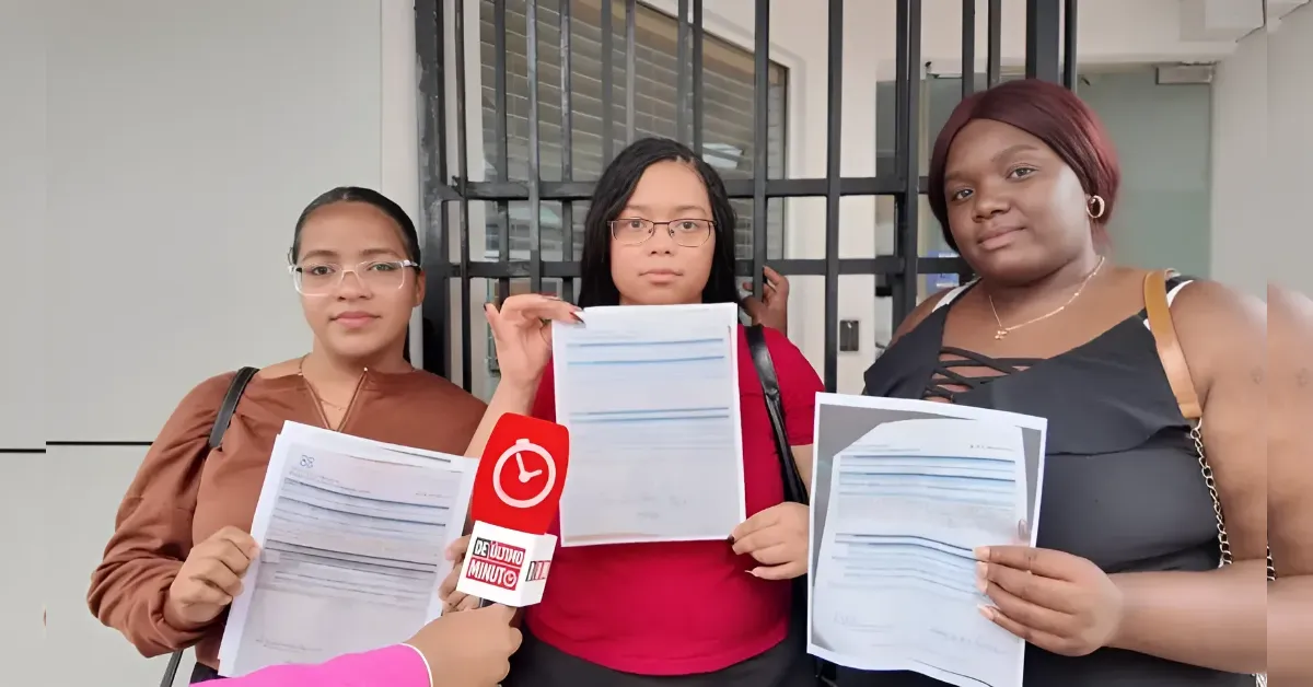 Three women stand side by side holding printed documents as a reporter with a red microphone interviews them. Their expressions are serious in a hall or lobby setting.