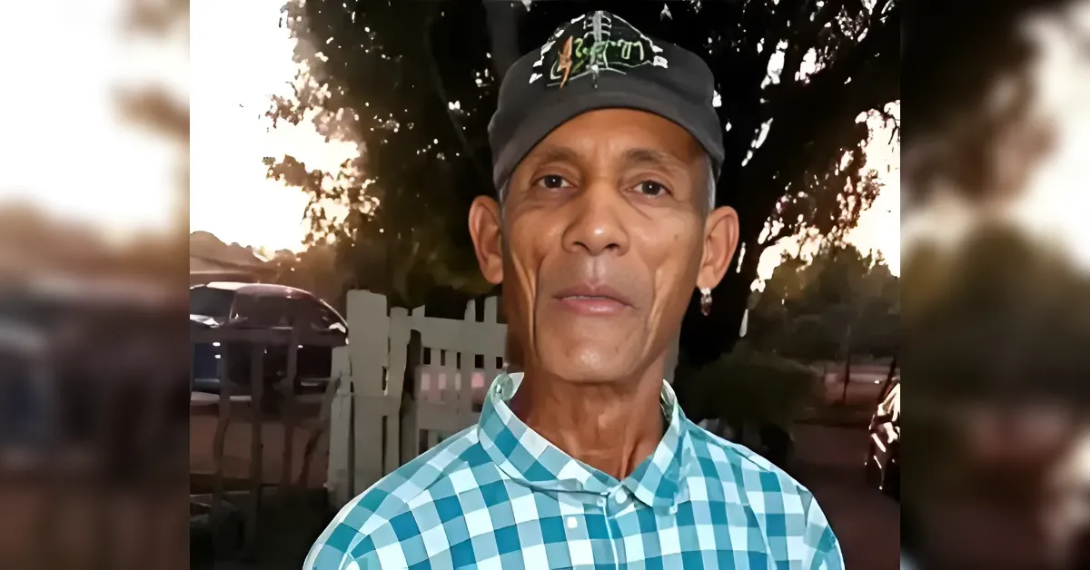 Older man wearing a teal and white checkered shirt and a dark cap, standing outdoors with a fence and trees in the background at sunset/ dusk.