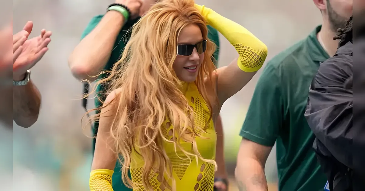 Woman in a bright yellow mesh outfit and sunglasses, adjusting her hair among people at a crowd event.