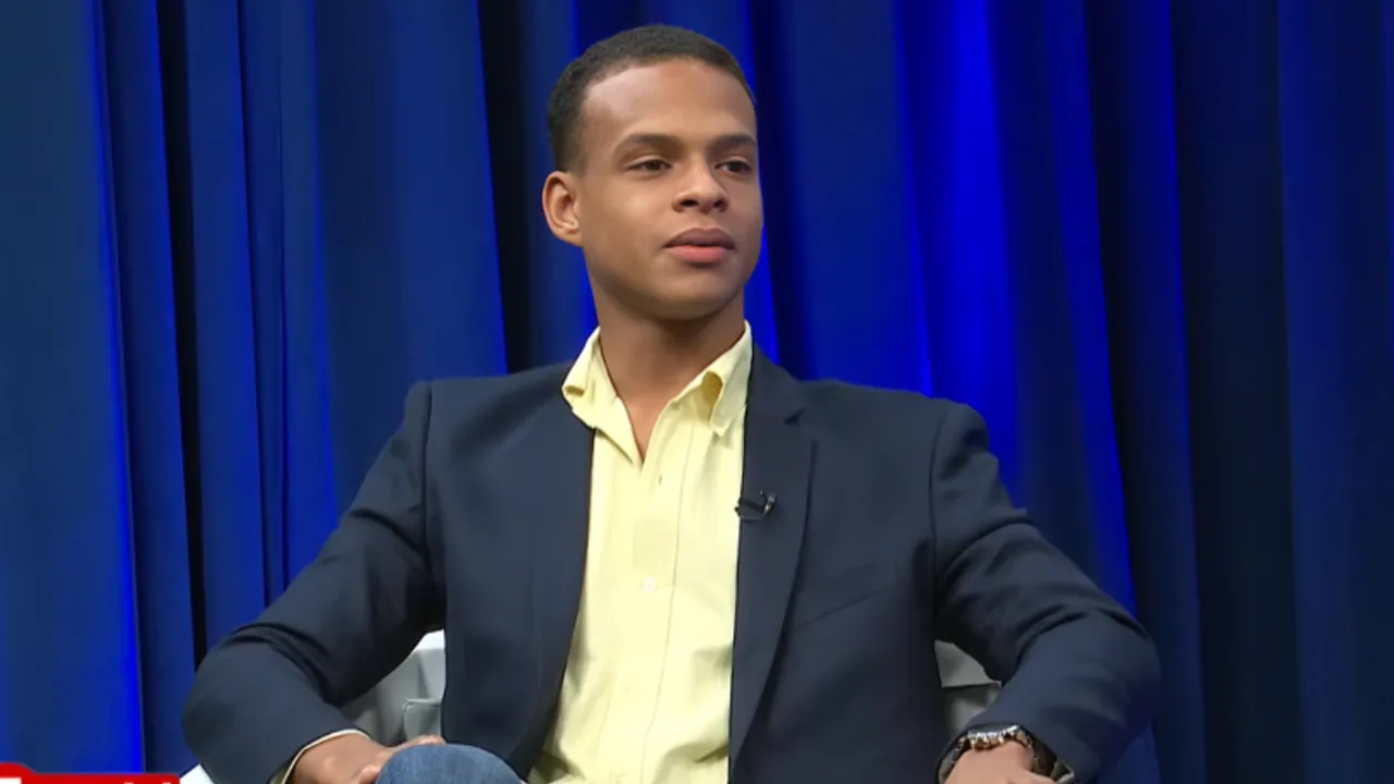 Young man in a navy blazer and pale yellow shirt sits on a stage with blue curtains, microphone clipped to his lapel.