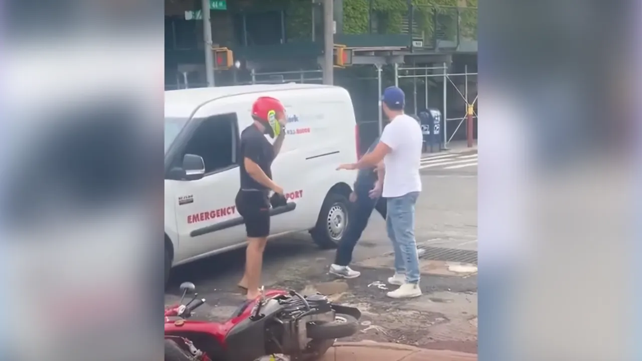 Two men stand near a white emergency transport van on a city street; one wearing a red helmet talks on a phone while the other gestures toward the sidewalk, with a red motorcycle on the ground nearby.