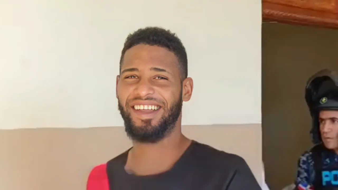 Smiling man with short hair and beard, wearing a dark shirt, against a beige wall; a person in a helmet appears in the doorway behind him.