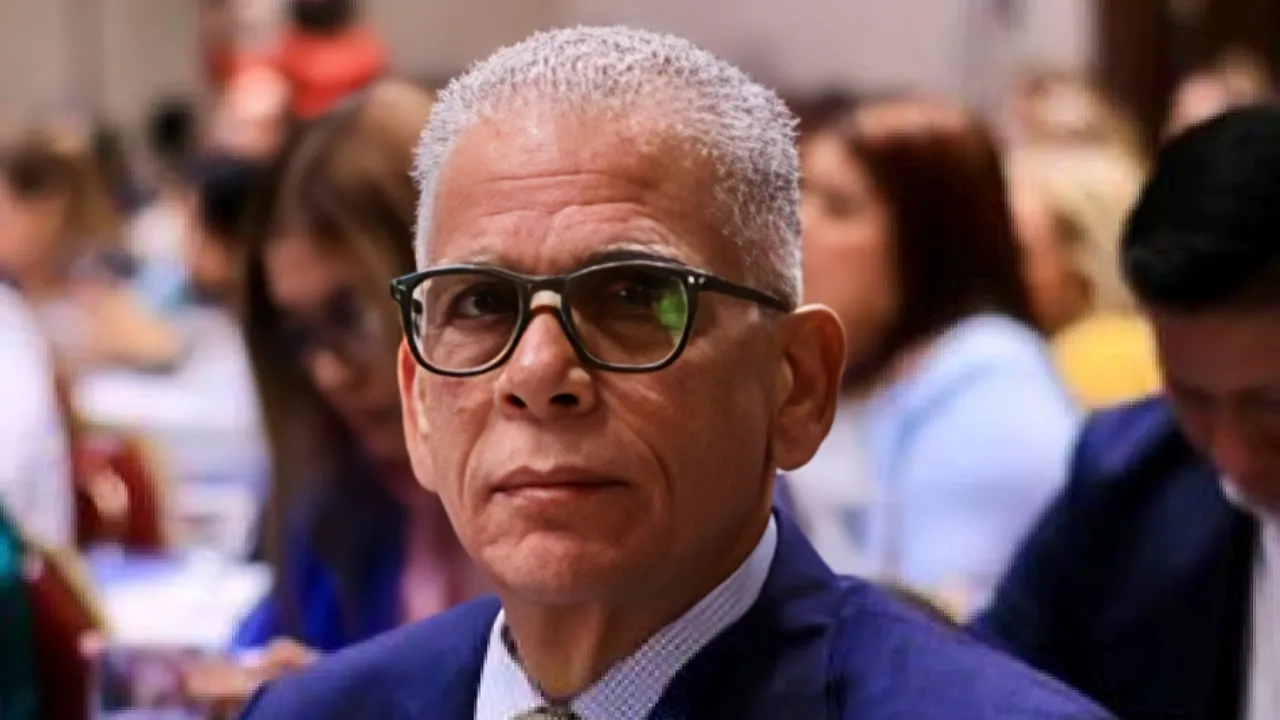 Portrait of a middle-aged man with short gray hair and black glasses, wearing a blue suit at a crowded formal event.