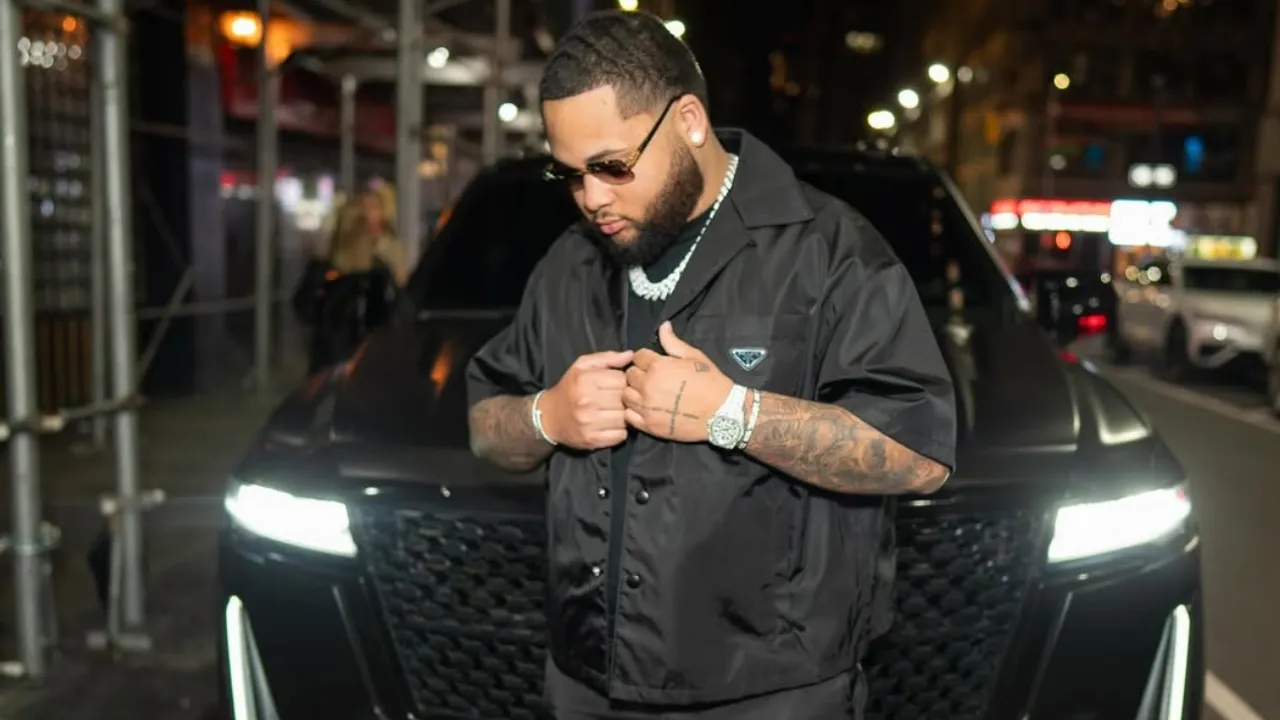 Man with beard and sunglasses stands in front of a black SUV at night, adjusting his black jacket amid city lights.