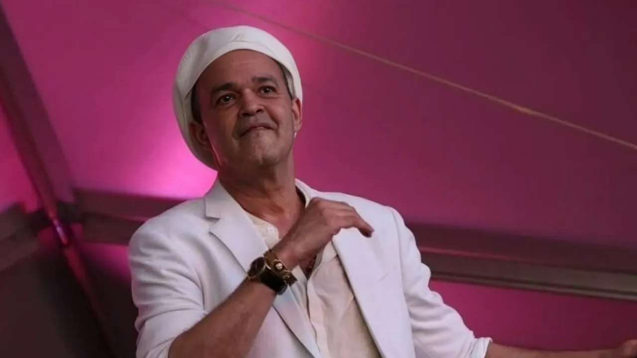 Man wearing a white suit and white turban, standing in a pink-lit room.