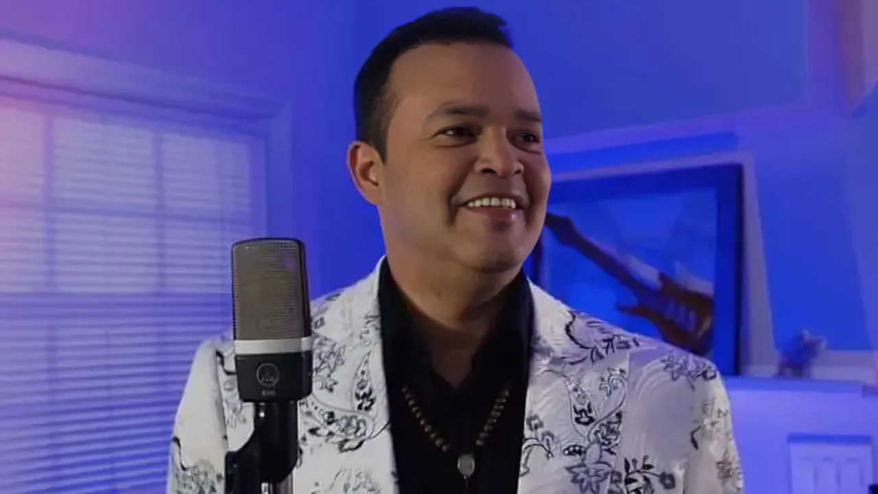 Man in a white floral blazer speaking into a studio microphone in a blue-lit room.