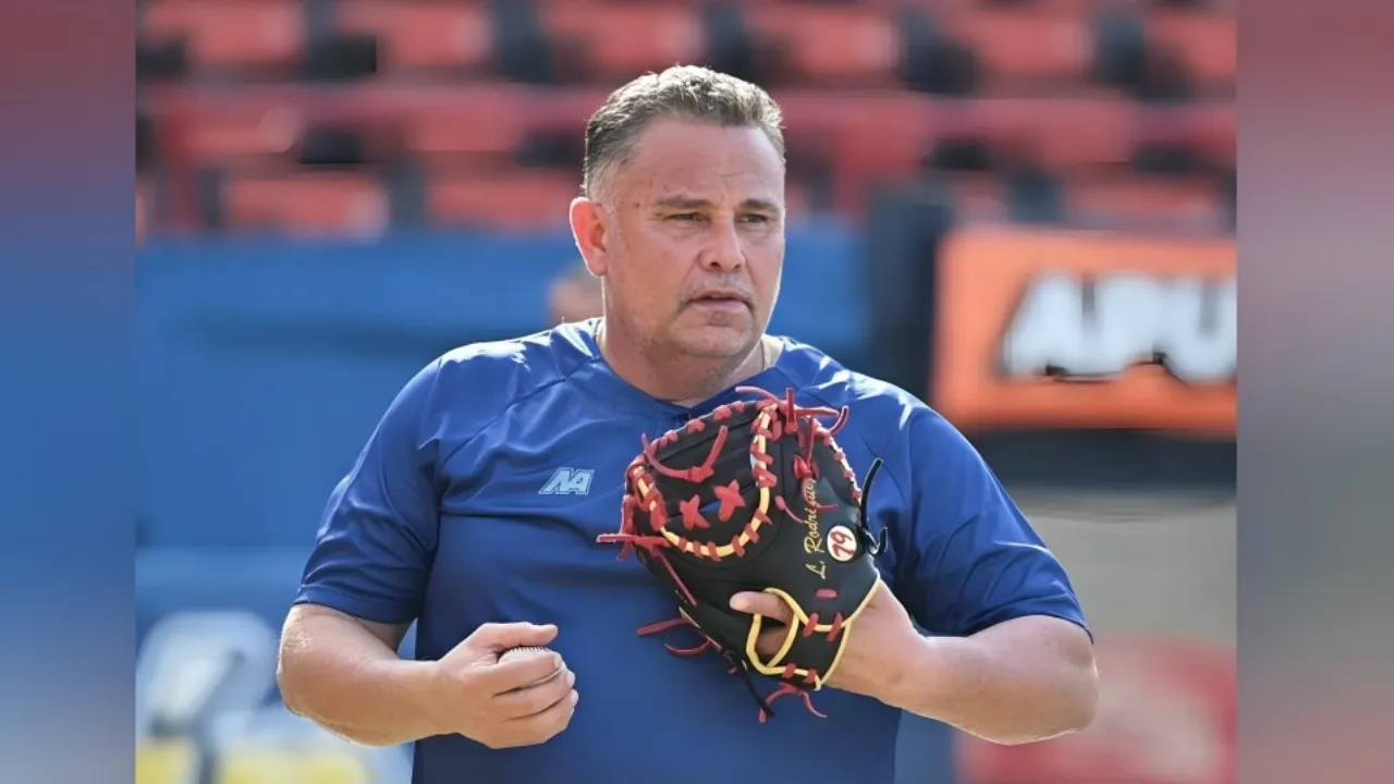 Man in a blue athletic shirt holding a baseball glove on his left hand, standing on a practice field with red stadium seating in the background.