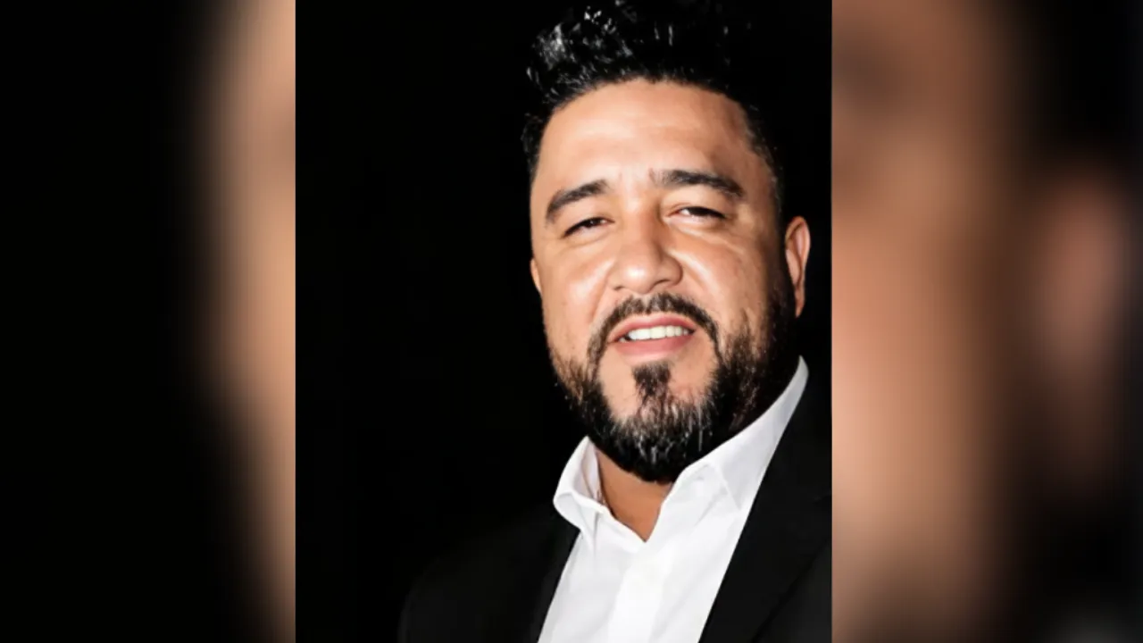 Man with dark hair and beard, wearing a black suit and white shirt, smiling at the camera against a dark background.