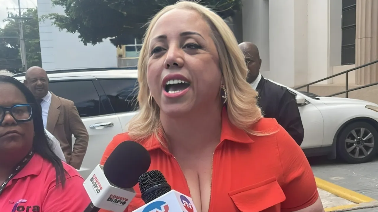 Woman in an orange blouse speaks to reporters outside a building, with multiple microphones in front of her.