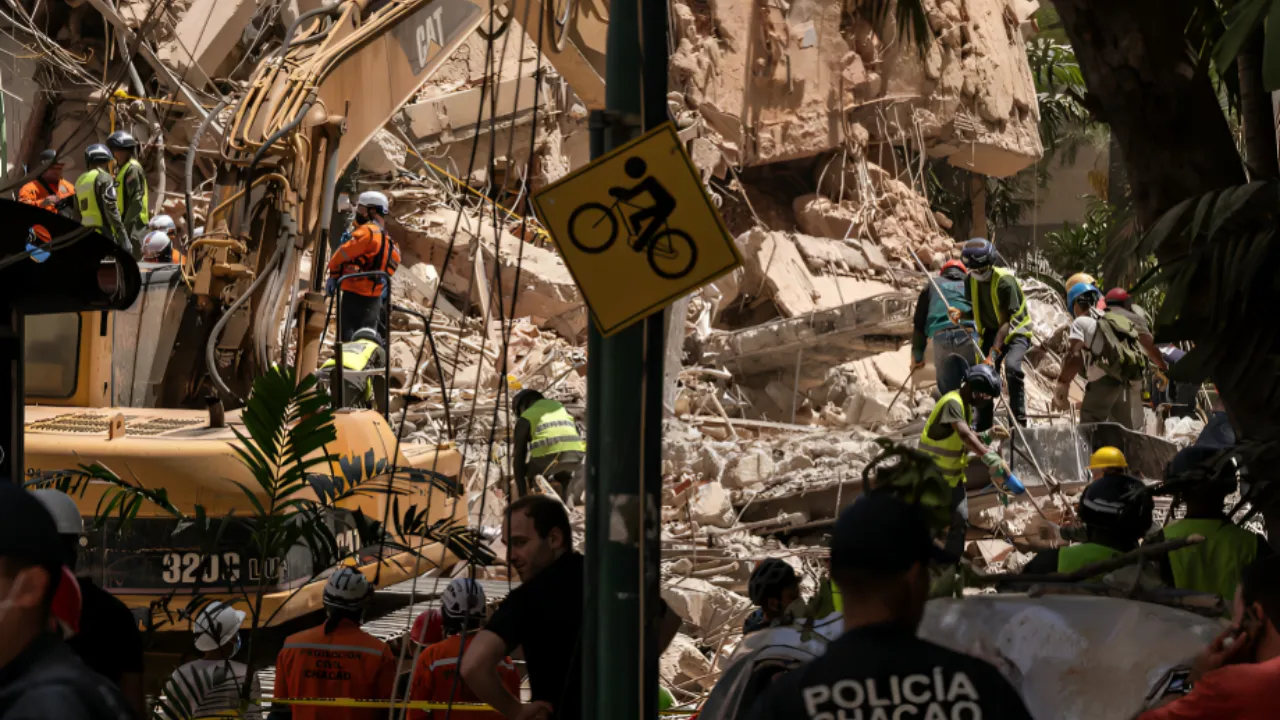 Rescue workers in helmets and reflective vests search through collapsed building rubble with a yellow excavator nearby.