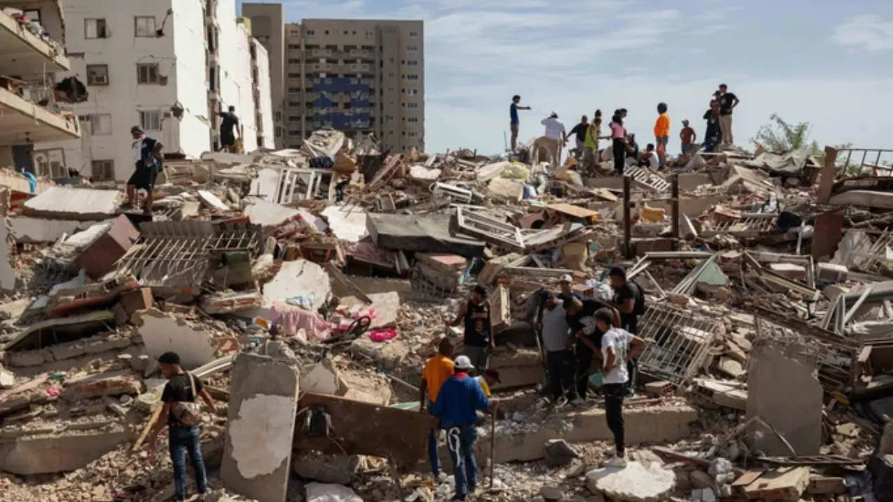 People stand and walk among rubble and collapsed buildings after a disaster, surveying the debris on a ruined urban landscape.