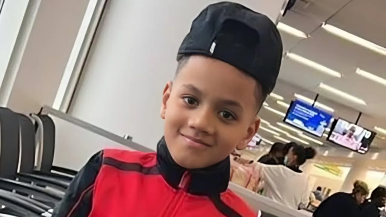 Smiling young boy in a red jacket and backward black cap sits in an airport terminal with seating and screens in the background.