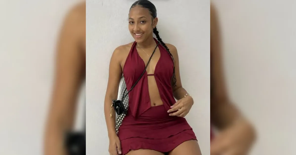 Smiling woman in a burgundy halter dress with a front cutout, sitting against a white wall with a small crossbody bag resting on her lap.
