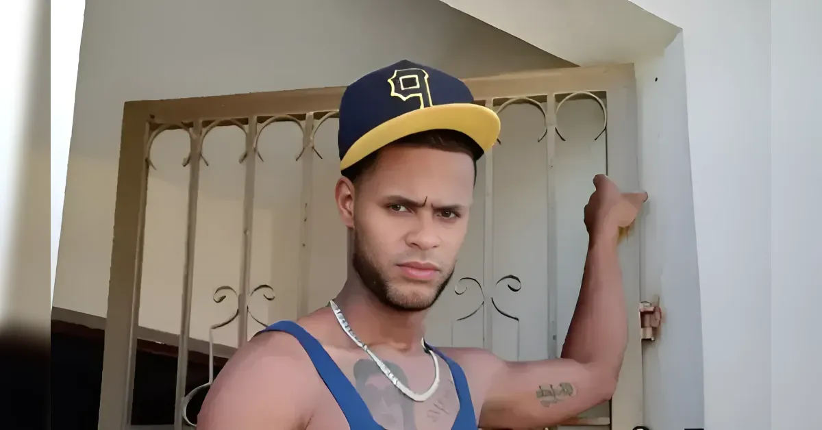 Young man in a blue tank top and two-tone yellow-and-navy cap leans on a metal gate, looking at the camera.