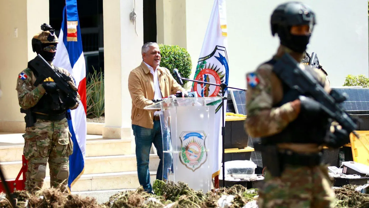 Man delivers a speech at an outdoor podium, flanked by two armed soldiers in camouflage gear; flags and a crest are visible behind him.