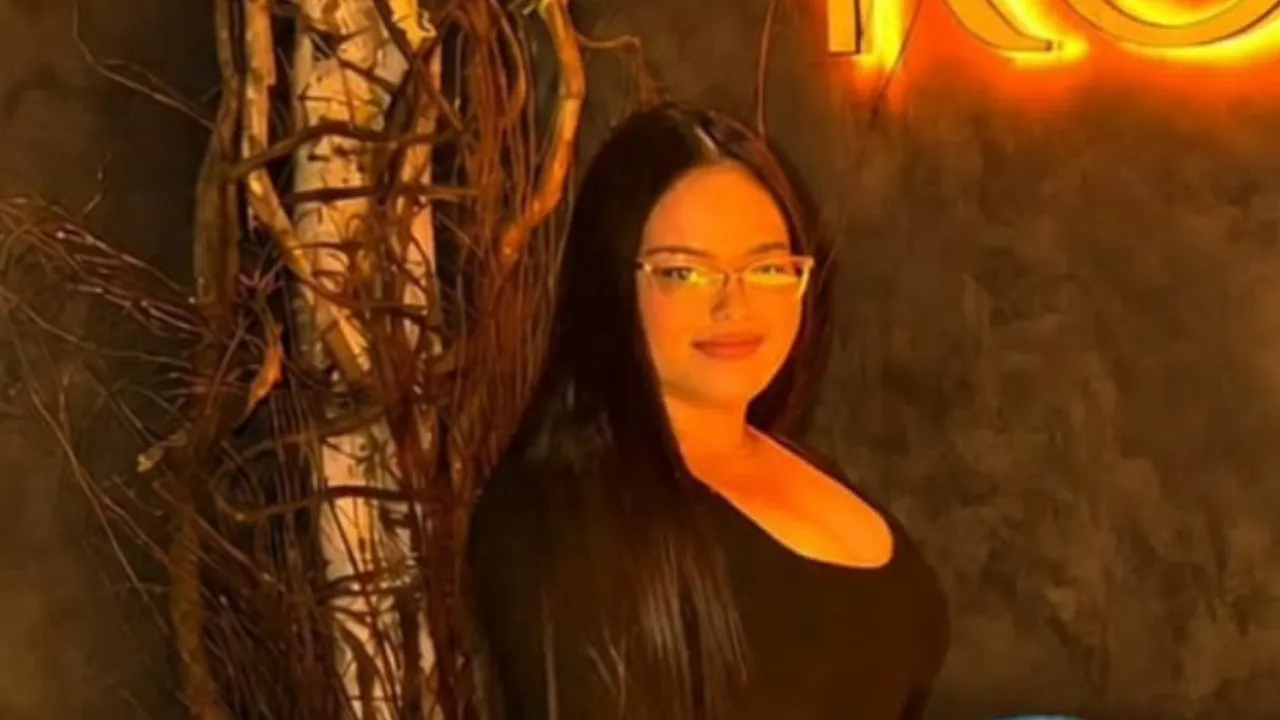 Woman with long dark hair and glasses standing against a vine-covered wall in a warm, orange-lit setting.
