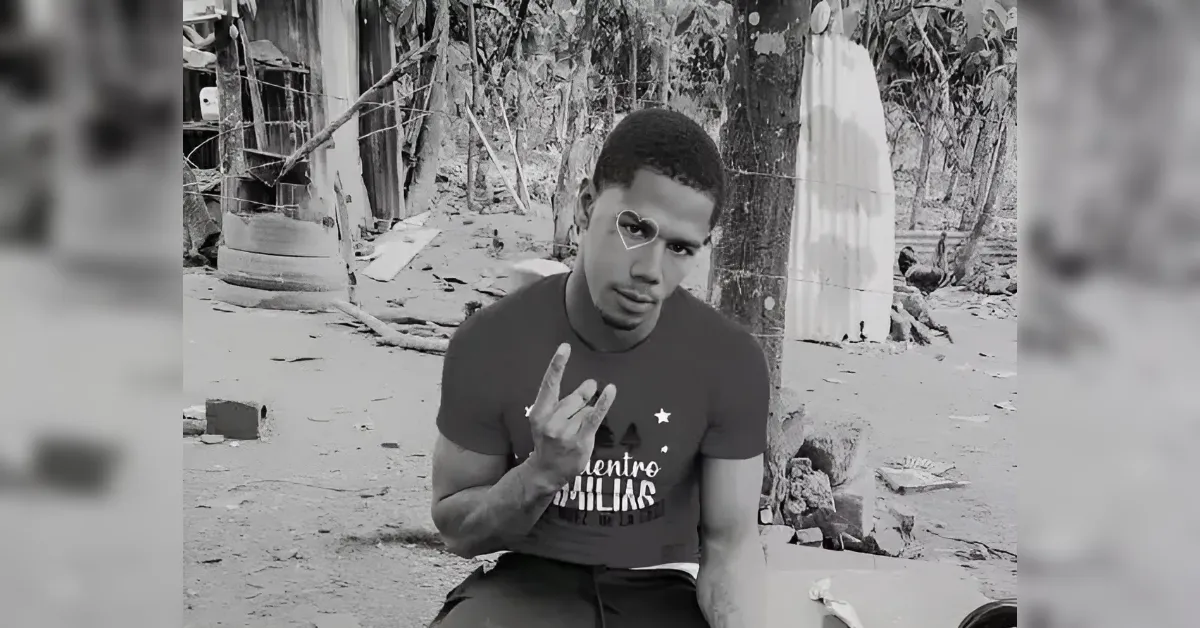Portrait of a young man sitting outdoors in a rural, grayscale scene, making a hand gesture toward the camera with a heart outline near his eye.