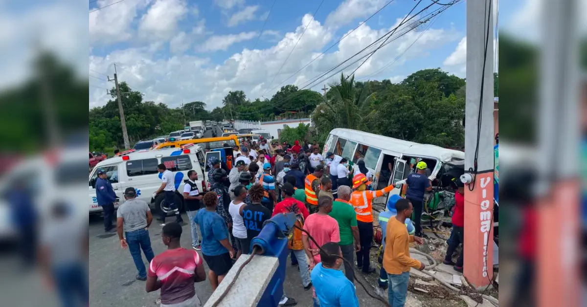 Crowded roadside after a bus crash with emergency responders and bystanders assisting at the scene. A white bus lies on its side amid debris and people in safety vests.