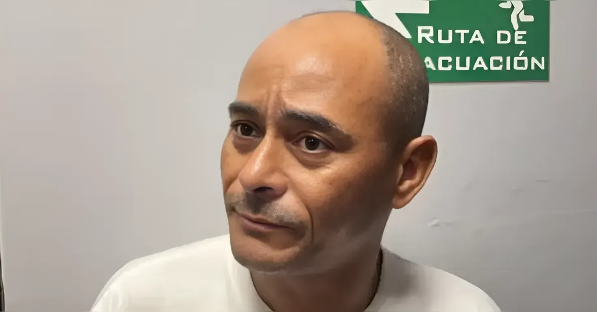 A man with a shaved head wearing a white shirt, looking to the side with a green evacuation sign in the background saying 'Ruta de evacuación'
