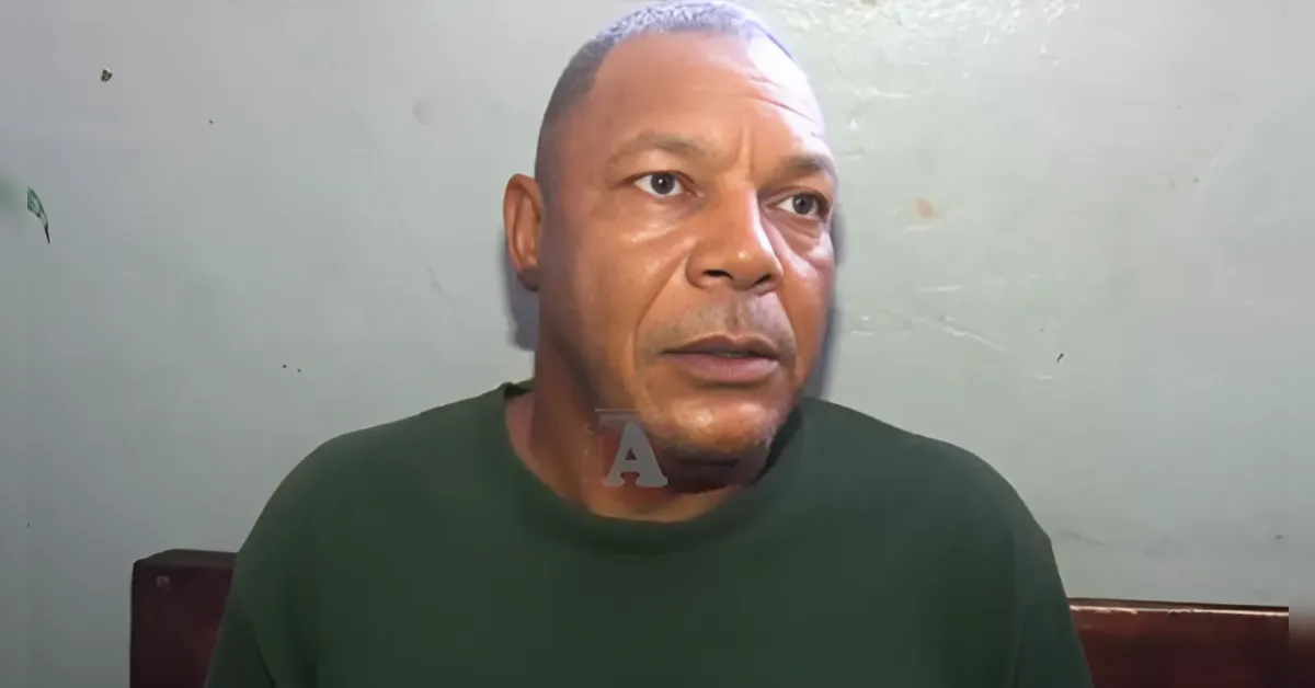 Close-up of a middle-aged man with a shaved head wearing a dark green shirt, seated against a plain gray wall.