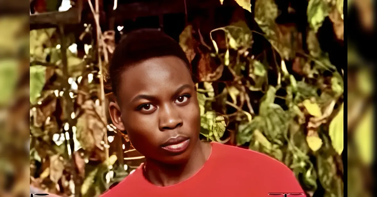Portrait of a young man with short hair wearing a red shirt, outdoors among green and brown leaves, looking at the camera.